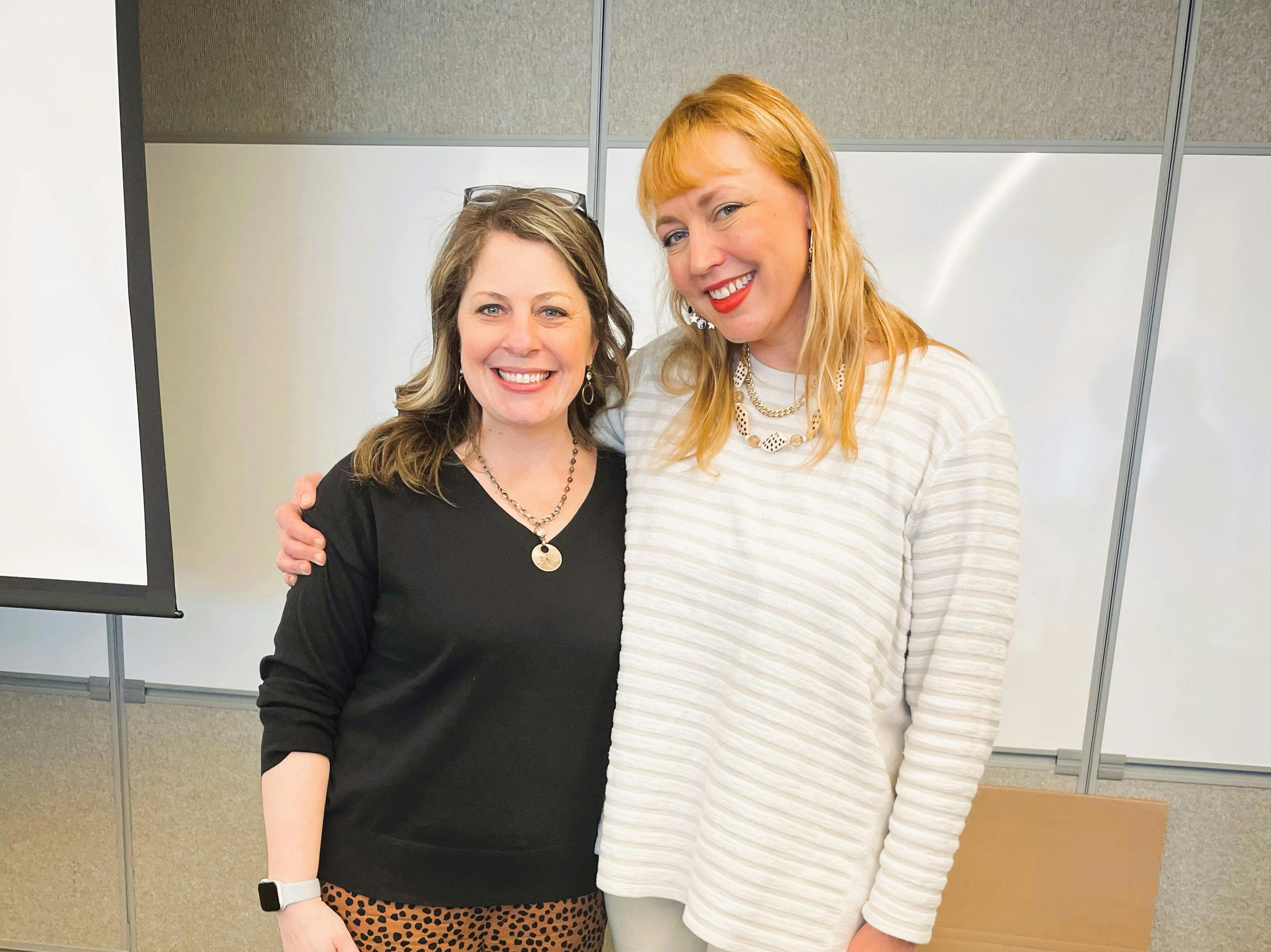 Two women side-hugging and smiling for the camera in front of a meeting-room whiteboard